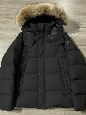  Canada Goose Wyndham  - Säljer en svart dunjacka från Canada Goose med stor fluffig päls på huvan och diskret logga på ärmen. Jackan har dragkedja och knappar framtill, två snedställda fickor och en quiltad design. Perfekt för kalla vinterdagar och riktigt snygg streetstil.