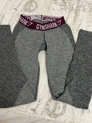 Grå Gymshark leggings med lila midjeband - Säljer ett par gråmelerade leggings från Gymshark med ett brett lila midjeband där det står GYMSHARK i vitt. De är tighta och har en hög midja, perfekta för träning. Materialet är stretchigt och mjukt för maximal komfort.