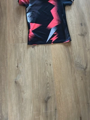 PSG x Jordan svart t-shirt - Säljer en svart PSG x Jordan t-shirt med feta röda och vita blixtmönster. T-shirten har korta ärmar, rund hals och är gjord i ett mjukt polyestermaterial. PSG-logga och Jumpman-logga broderade på bröstet. Perfekt för dig som älskar fotboll och streetwear.