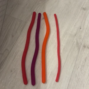 Monkey nudels - Detta är fyra färgglada, böjbara silikonstavar i rött, lila och orange. De är flexibla och kan användas som sensoriska leksaker eller stressleksaker. Alla stavar är i gott skick utan synligt slitage. Perfekt för barn eller vuxna som gillar taktila produkter.