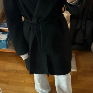 Svart kappa med bälte - Säljer en svart kappa med bälte i midjan och klassisk krage. Jackan är långärmad och har en stilren, enkel design som funkar till många olika outfits. Materialet känns mjukt och värmande, perfekt för kyligare dagar. Jag är ca 163 o den går till knäna på mig. Varm o skön.