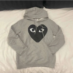 Grå hoodie Comme des Garçons Play - Grå hoodie från Comme des Garçons Play med stort svart hjärta och ögon på bröstet. Klassisk huva med snörning och magficka. Mjuk bomullskänsla och ribbade muddar vid ärmslut och nederkant. Perfekt för en chill och stilren look.