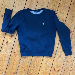 Mörkblå sweatshirt från Polo Ralph Lauren - Mörkblå sweatshirt från Polo Ralph Lauren med klassisk rund hals och broderad logga på bröstet. Tröjan har långa ärmar och ribbade muddar vid ärmslut och nederkant. Perfekt för en chill och stilren look. Liten i storleken, upplevs mer som en XS.