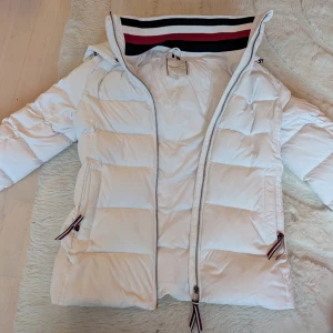 Vit pufferjacka från Tommy Hilfiger - Säljer en snygg vit pufferjacka från Tommy Hilfiger med huva och dragkedja. Jackan har broderad logga på ärmen, randiga detaljer i rött, vitt och svart samt två fickor med dragkedja. Perfekt för kalla dagar och riktigt stilren look.