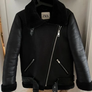 Svart pälsjacka från Zara med bälte - Snygg svart pälsjacka från Zara med bred krage, dragkedja i sned modell och två fickor med dragkedjor. Jackan har bälte i midjan och mjukt foder, perfekt för kalla dagar. Stilsäker och cool look med detaljer i skinnimitation.