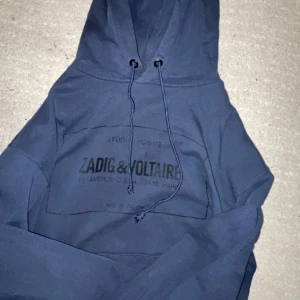 Mörkblå hoodie från Zadig & Voltaire - Säljer en mörkblå hoodie från Zadig & Voltaire med tryck framtill och coola snören i huvan. Tröjan har klassisk känguruficka och långa ärmar. Perfekt för dig som gillar streetstyle och vill ha något bekvämt och snyggt.