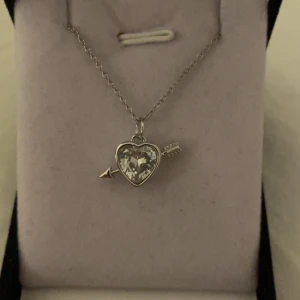 Halsband med hjärthänge och pil silver vit - Snyggt silverfärgat halsband med ett gnistrande hjärthänge i glas eller kristall, dekorerat med en pil som går genom hjärtat. Perfekt accessoar för dig som gillar romantiska detaljer och vill sticka ut lite extra. Rostfri 