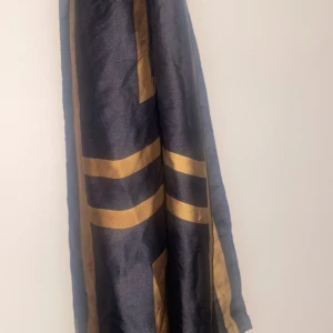 Svart och guldig halsduk i silke - Materialet är silkeslent och ger en lyxig känsla. Perfekt att svepa runt halsen för att lyfta din outfit med lite extra edge. Mönstret är unikt! Aldrig använd. 