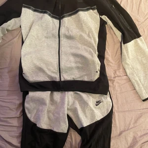 Nike hoodie i svart och grå med dragkedja - Nike hoodie i svart och ljusgrått med hel dragkedja och huva. Snygg sportig look med svart Nike-logga på bröstet och ärm med liten ficka. Tillverkad i mjuk bomullsblandning, perfekt för träning eller chill. Matchande byxor ingår. Bra skick använts några gånger. Tröja och byxor måste man köpa tillsammans kan inte bara köpa en produkt. Inte äkta 