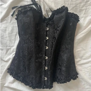 Svart korsett med snörning och spets - Säljer en svart korsett med snyggt broderat mönster och volangkant. Framsidan har metallknäppning och baksidan har snörning för perfekt passform. Materialet är 90% polyester och 10% elastan, vilket gör den både bekväm och formande. Perfekt för att lyfta din outfit!