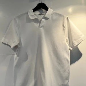 Vit pikétröja från Bläck - Klassisk vit pikétröja från Bläck i storlek S. Tröjan har korta ärmar, mjuk krage och en stilren design utan knappar. Perfekt för en clean och fräsch look. Tillverkad i bomull för en skön känsla hela dagen.