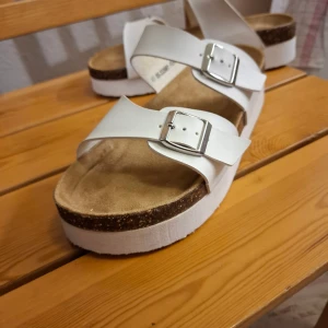 Vita sandaler med plattform och spännen - Säljer ett par vita sandaler med breda remmar i syntet och två silvriga spännen. Sandalerna har en plattformsula med korkdetalj och mjuk innersula i beige. Perfekta för dig som gillar chunky stil och vill ha något som sticker ut. Ca 3 cm sula. Fotbädd ca 26 cm.