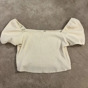 Ribbad offshoulder topp från H&M - Säljer en ljusbeige ribbad topp från H&M i storlek M. Modellen är offshoulder med puffiga korta ärmar och croppad passform. Materialet är mjukt och stretchigt, perfekt för att skapa en trendig look. Supersnygg att styla med jeans eller kjol!
