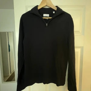 Svart merinoullströja från These Glory Days - Svart långärmad tröja i 100% merinoull från These Glory Days. Tröjan har en stilren half zip-dragkedja vid kragen och en klassisk passform. Perfekt för dig som gillar minimalistisk och tidlös stil.