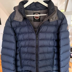 Mörkblå dunjacka från Colmar - Snygg marin blår pufferjacka från Colmar med hög krage och dragkedja framtill. Jackan har ett stilrent, quiltat mönster och Colmar-logga på ärmen. Perfekt för kalla dagar och riktigt skön att bära. Size 50 och några små skrapmärken på vänsterarmen men syns knappt då jacka är mörk. 