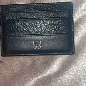 Svart kortfodral från Hugo - Snyggt svart kortfodral från Hugo i äkta skinn. Stilren design med både slät och präglad yta, flera kortfack och diskret logga framtill. Perfekt för dig som vill ha ett smidigt och modernt sätt att bära dina kort.