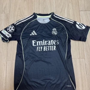 Real Madrid Mbappé fotbollströja M - Snygg mörkblå Real Madrid fotbollströja från Adidas med Mbappé och nummer 10 på ryggen. Tröjan har korta ärmar, silverfärgade detaljer och klubbmärke på bröstet. Tillverkad i lätt och ventilerande material med AEROREADY-teknologi.