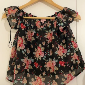 Blommig offshoulder blus - Säljer en luftig och söt blommig blus med offshoulder-modell. Toppen är svart med färgglada blommor i rosa, rött och grönt. Den har korta, volangiga ärmar och är gjord i ett tunt, transparent material som känns riktigt somrigt.