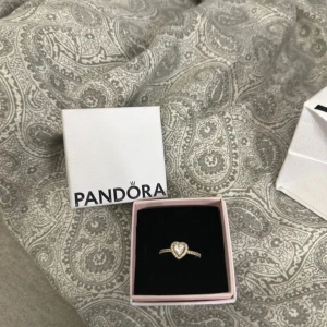Pandora ring med hjärta i guld - Snygg ring från Pandora i guld med ett glittrigt hjärta som mittpunkt. Ringen har små gnistrande stenar längs bandet och levereras i original ask. Perfekt för dig som vill ha något extra på fingret. storlek 52, perfekt till julklapp