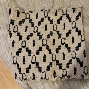 Beige och svart mönstrad topp - Säljer en beige topp med svart grafiskt mönster. Toppen har en smockad, elastisk struktur som ger en tight passform och volangkant upptill och nedtill. Perfekt för dig som gillar unika detaljer och vill sticka ut med ett coolt mönster.