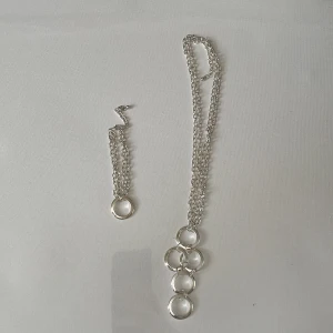 Set med silverfärgat halsband och armband - Säljer ett snyggt set med halsband och armband i silverfärg. Båda har grova kedjor och stora, blanka ringar som detaljer. Halsbandet har flera ringar i en rad, medan armbandet har en stor ring i mitten. Perfekt för dig som gillar statement-smycken med modern känsla.