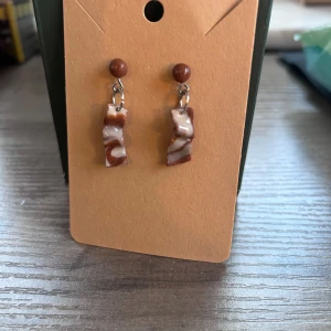 Bruna och vita örhängen med marmorlook - Unika örhängen med bruna och vita marmorliknande hängen i oregelbunden form. Fästs med en brun kula upptill och har coolt, konstnärligt uttryck. Perfekta för dig som vill sticka ut med accessoarerna.