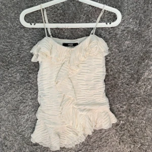 Vit volangig linnetopp från Bik Bok - Supersöt vit linnetopp från Bik Bok med tunna axelband och volangdetaljer framtill och nedtill. Toppen har ett strukturerat tyg med vågigt mönster och är perfekt för dig som gillar en feminin och trendig look. Sitter snyggt och är lätt att matcha med jeans eller kjol.