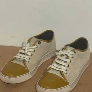 Lanvin sneakers i beige mocka och gult lack - Snygga sneakers från Lanvin i beige mocka med detaljer i gul lack på tå, häl och runt öppningen. Skorna har vita snören och en klassisk platt sula. Perfekta för dig som vill ha en unik och stilren look med lyxig känsla.