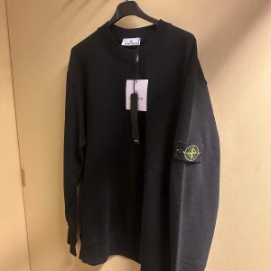 Svart sweatshirt,  Stone Island. - Svart sweatshirt från Stone Island med klassisk rund hals och lång ärm. Ikonisk logotyp-patch på vänster ärm. Tröjan har en loose passform och är tillverkad i mjuk bomull, perfekt för chill dagar. Enkelt och stilrent plagg som lyfter din streetwear-look. 