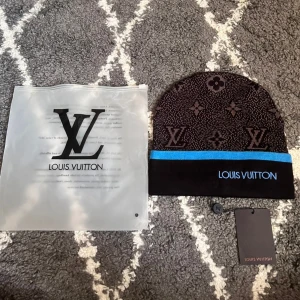 L-mössa -  Exklusiv mössa från L i  grått och svart med klassiskt L-monogram och blommönster. Mössan har en svart uppvikt kant med blå rand och broderad L-logga framtill. Tillverkad i mjukt stickat material, perfekt för kyliga dagar.