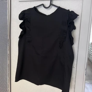 Svart Top med volangärmar - Säljer en svart Top  med rund hals och snygga volangdetaljer på axlarna. Toppen är ärmlös och har en enkel knapp i nacken. Perfekt för dig som gillar stilrena plagg med en twist.