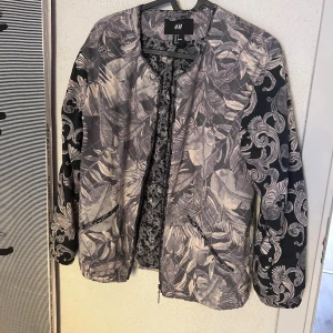Quiltad mönstrad jacka från H&M - Quiltad jacka från H&M i storlek 34 med grått och svart bladmönster och detaljerade mönstrade ärmar. Jackan har dragkedja framtill, två fickor och rund hals. Materialet är lätt, bomull och polyester, perfekt som kavaj över klänning eller Top, eller lager-på-lager under höst eller vår.
