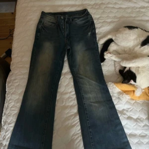 Blå bootcut jeans med broderi - Säljer ett par blå bootcut jeans med snyggt broderade detaljer på bakfickorna. Jeansen har slitningar och tvättade partier för en cool look, samt fickor med lock och knapp. Perfekt för dig som gillar unik stil.