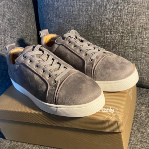 Grå mocka sneakers från Louboutin - Snygga grå sneakers från Christian Louboutin i mjuk mocka med vit platt sula och klassisk snörning. Insidan är lyxigt guldfärgad och yttersulan har den ikoniska röda Louboutin-signaturen. Perfekt för dig som vill sticka ut med exklusiv stil.