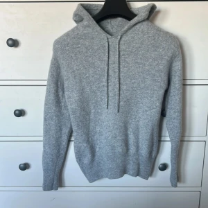 Grå stickad hoodie i kashmir - Mysig grå hoodie i kashmir med huva och snörning. Tröjan är stickad och har långa ärmar samt ribbade muddar vid ärmslut och nederkant. Perfekt för kyliga dagar och enkel att matcha med jeans eller mjukisbyxor.