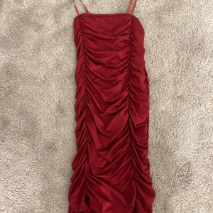 Röd långklänning med rynk från SHEIN - Snygg röd långklänning från SHEIN med smala axelband och rynkade detaljer längs hela klänningen. Figurnära passform och tillverkad i ett mjukt, glansigt polyestertyg som ger en lyxig känsla. Perfekt för dig som vill sticka ut. Använt klänningen endast 2 gånger.