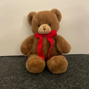 GUND Teddy Bear - Mjuk och klassisk GUND nallebjörn med röd rosett. Maskintvättbar och i mycket fint skick, utan synligt slitage eller defekter. Perfekt som present eller mysig dekoration. Pälsen är mjuk och tät, och nallen har en söt, vänlig look.
