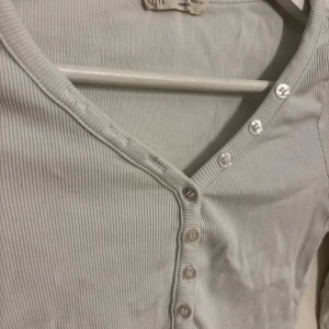 Vit ribbad långärmad topp - Säljer en vit ribbad långärmad topp. Toppen har v-ringning och knappar framtill som ger en snygg detalj. Materialet är mjukt och stretchigt, perfekt för en chill och stilren look. Passformen är slim och toppen är något croppad.