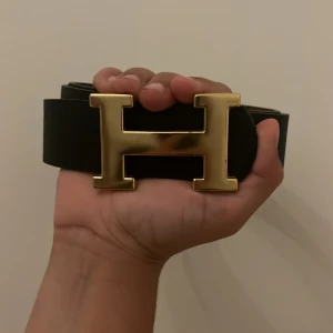 Svart H bälte med guldspänne - Säljer ett svart bälte från H med ett stort, ikoniskt H-spänne i guld. Bältet är stilrent och tillverkat i skinn, perfekt för att lyfta din outfit med en lyxig touch. Passar dig som gillar klassiska accessoarer med statement-detaljer.