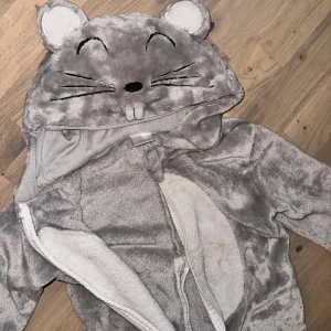 Grå dräkt Grå mjuk fleeceoverall med musmotiv - Supergosig grå overall i fleece med huva som har söta musöron, broderat ansikte och tänder. Dragkedja framtill och vita detaljer på magen och öronen. Perfekt för kyliga dagar när man vill vara både varm och gullig.Söt grå musdräkt med huva. Dräkten har en dragkedja framtill och en vit mage. Huvans design har broderade detaljer som ögon, morrhår och små tänder, samt små runda öron upptill. Mouse 🐭 