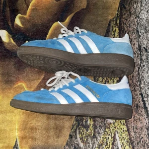 Adidas Spezial blå sneakers mocka - Säljer ett par Adidas Spezial sneakers i ljusblå mocka med vita detaljer och klassiska tre ränder på sidan. Skorna har bruna gummisulor, vit snörning och rund tå. Adidas-logga och 'Special' i guld på sidan. Perfekt för dig som gillar retrostil och streetwear.