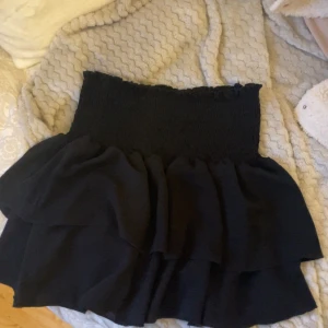 Svart volangkjol med smock - Supersöt svart kortkjol med bred smock i midjan och dubbla volanger som ger en flowy känsla. Kjolen är enkel att matcha och har en feminin siluett. Perfekt för dig som gillar en trendig och bekväm look.