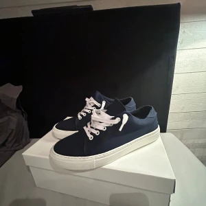 Mörkblå sneakers med vit sula - Snygga mörkblå sneakers med vit platt sula och vita snören. Skorna har en klassisk rund tå och är tillverkade i textilmaterial. Perfekta för dig som gillar stilrena och enkla sneakers med kontrast mellan färgerna.