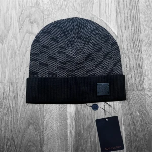 Svart rutig mössa  - Exklusiv svart mössa från  med klassiskt rutigt mönster och ribbad uppvikt kant. Märkets logopatch i svart skinn framtill ger en lyxig touch. Materialet är mjukt och stickat, perfekt för att hålla dig varm och stilren.