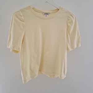 Ljusgul topp med puffärm från Monki - Supersöt ljusgul topp från Monki i 100% bomull. Modellen har korta puffärmar och rund halsringning, vilket ger en trendig och lekfull vibe. Perfekt att styla med jeans eller kjol för en fräsch look.