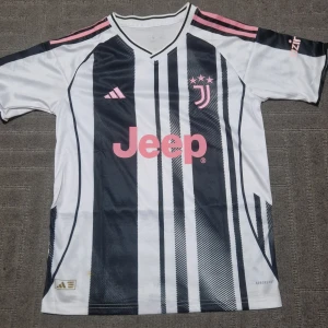Juventus fotbollströja Adidas svartvit - Juventus fotbollströja från Adidas med klassiska svarta och vita ränder, rosa detaljer och logga. Tröjan har rosa Adidas-ränder på axlarna, Jeep-tryck på bröstet och Juventus klubbmärke. Tillverkad i lätt och ventilerande material.