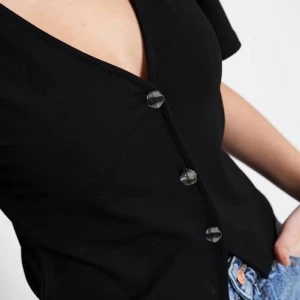 Svart kortärmad blus med knappar - Svart blus med korta ärmar och v-ringning. Toppen har tre dekorativa knappar framtill och är croppad i modellen. Perfekt att matcha med jeans för en chill look. Materialet känns mjukt och lätt, troligtvis bomull eller polyester.