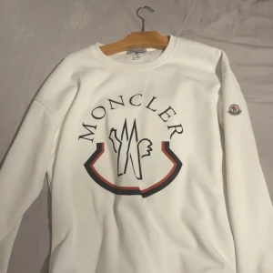 Vit sweatshirt från Moncler - Snygg vit sweatshirt från Moncler med stort tryck på bröstet och klassisk logga på ärmen. Tröjan har rund halsringning och långa ärmar. Perfekt för dig som gillar stilrena och exklusiva plagg. Storleken XXL 75kg -90 kg. Passar XL
