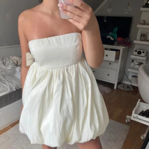 Vit axelbandslös kortklänning från Malina - Superfin vit axelbandslös kortklänning från Malina med ballongformad kjol och slät överdel. Klänningen har en lyxig satinliknande finish och är perfekt för dig som vill sticka ut med en unik siluett. Passar dig som gillar trendiga och eleganta plagg. Helt oanvänd med prislappen kvar. Perfekt klänning till studenten!😍