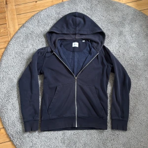 Mörkblå hoodie från Jack & Jones - Mörkblå hoodie från Jack & Jones i storlek XS. Klassisk modell med dragkedja framtill, två fickor och stor huva. Tillverkad i mjuk bomullsmix med fleecefoder på insidan för extra komfort. Perfekt för kyliga dagar och enkel att matcha med jeans eller joggers.